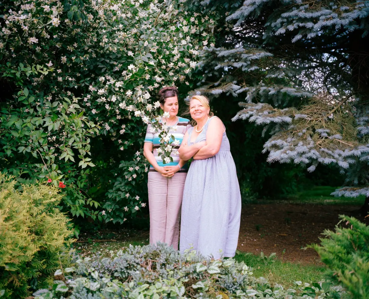 Two adults pose together in a garden, standing among white flowering shrubs and blue spruce branches.