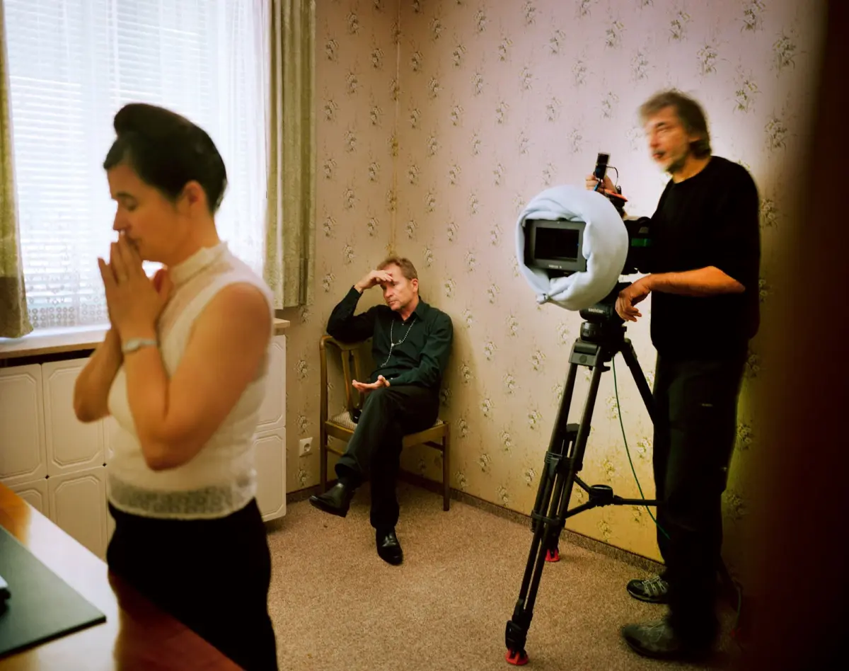 Film crew in a floral wallpapered room; man operates a tripod-mounted video camera; another man sits with hand on brow; woman prays in foreground.