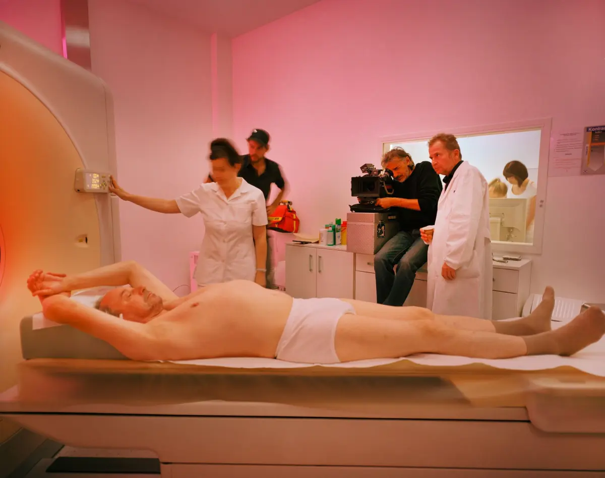 Adult patient lying on MRI scanner table; camera operator and clinicians in pink-lit radiology room.