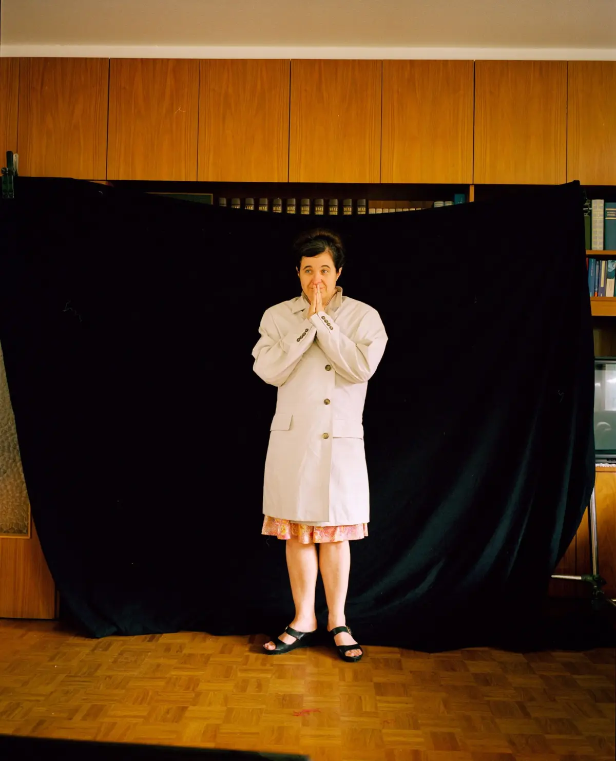 Person wearing a beige trench coat stands with hands pressed together in front of a black backdrop, wooden cabinets above.