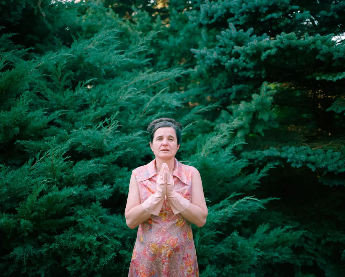 Person in a pink floral dress with cream gloves, hands pressed together in front of dense evergreen shrubs outdoors.