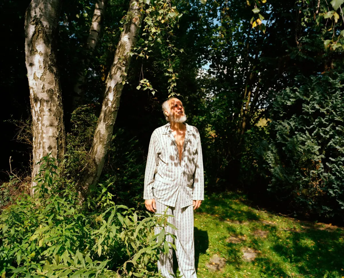 Elderly person with white beard stands among birch trees, gazing upward at hanging branches in a sunlit forest.