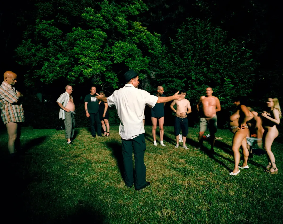 Adult in white shirt with outstretched arms addresses a circle of shirtless adults outdoors at night, with dense green trees in the background.