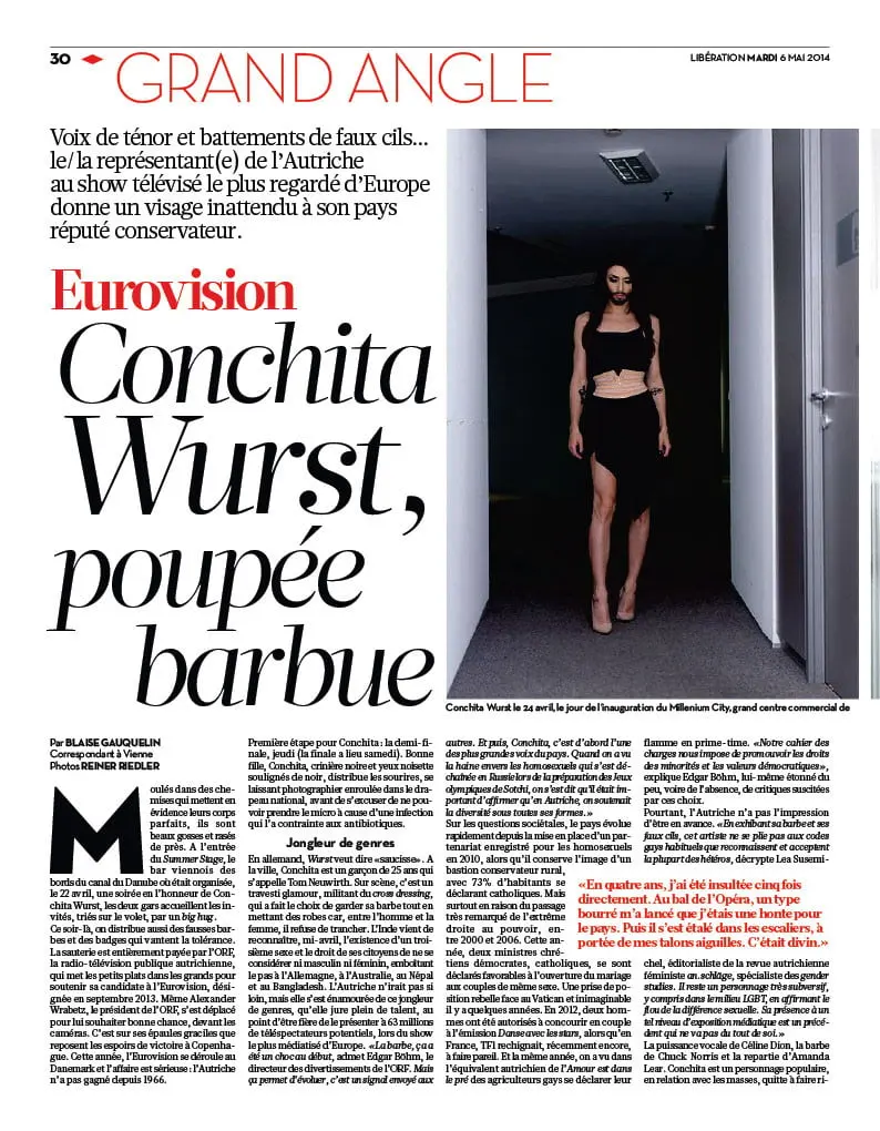 Bearded drag performer in a black crop top and long skirt stands in a doorway in a dim hallway.