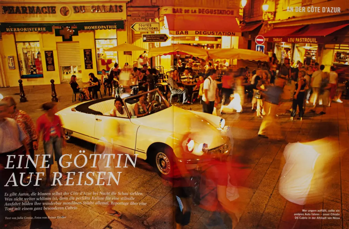 White vintage convertible parked in front of a crowded outdoor café on a European street at dusk.