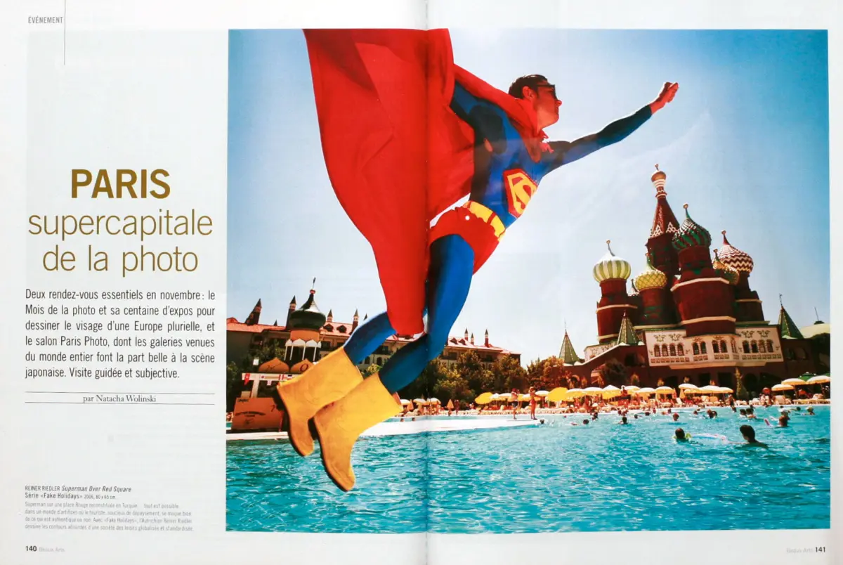 Superhero in blue suit, red cape, and yellow boots leaps over a bright pool; colorful onion-domed architecture rises in the background.