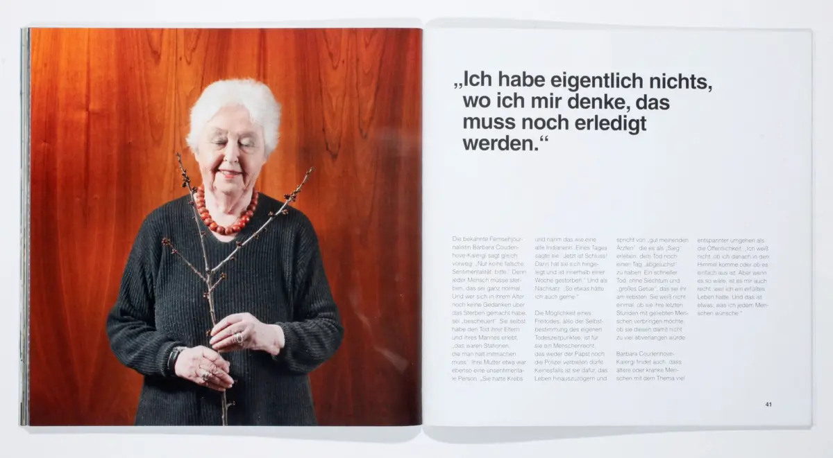 Elderly woman with white hair, dark sweater, red bead necklace, holding a budding branch; wood panel background; adjacent page with German quote.