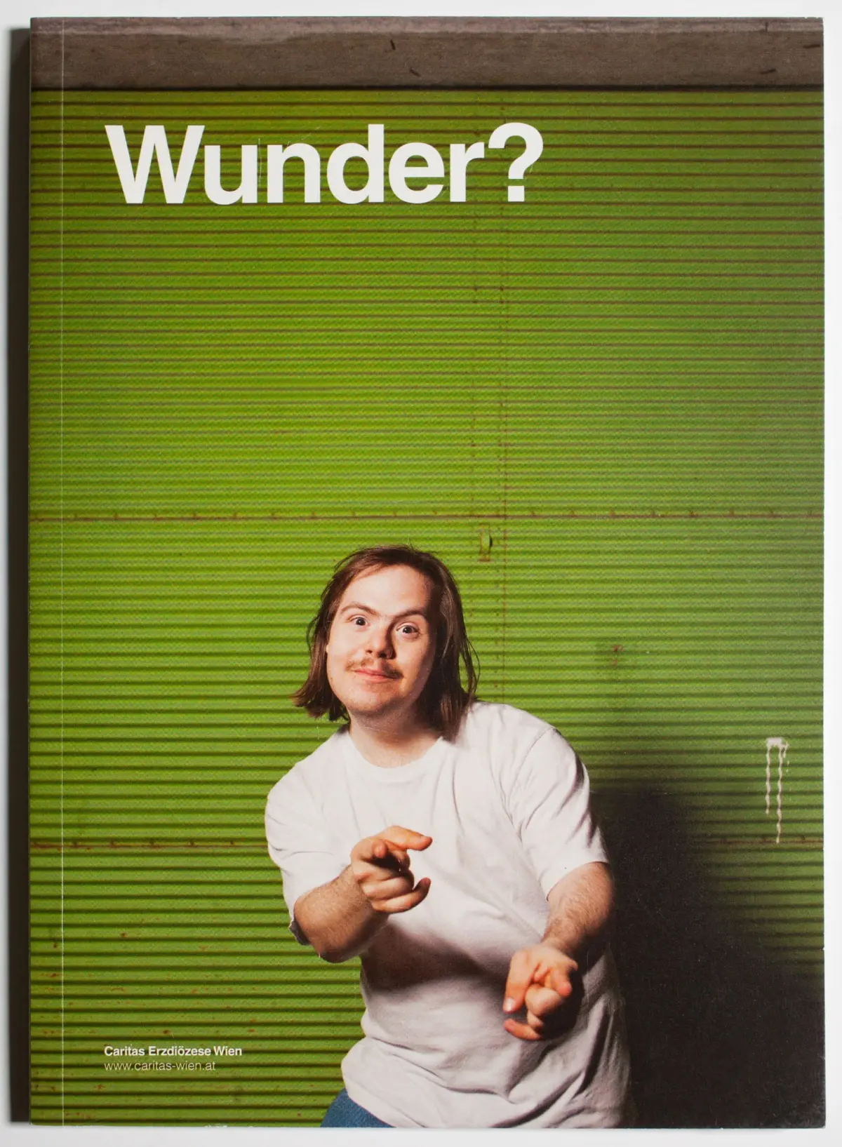 Person with shoulder-length brown hair in a white T-shirt gestures toward the camera against a green corrugated metal backdrop; Wunder? text at top.