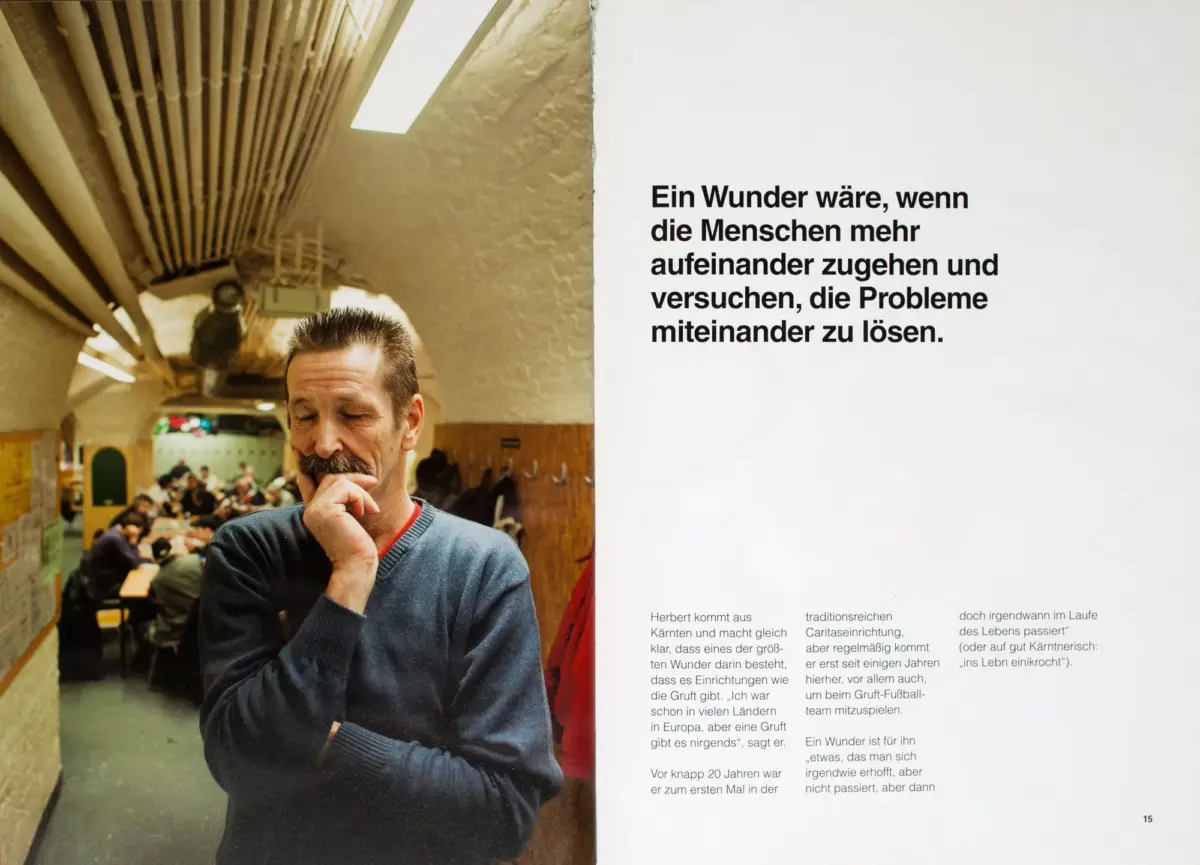 Adult person with short hair and mustache, chin on hand, in a busy classroom-like hall with long tables and wooden ceiling beams.