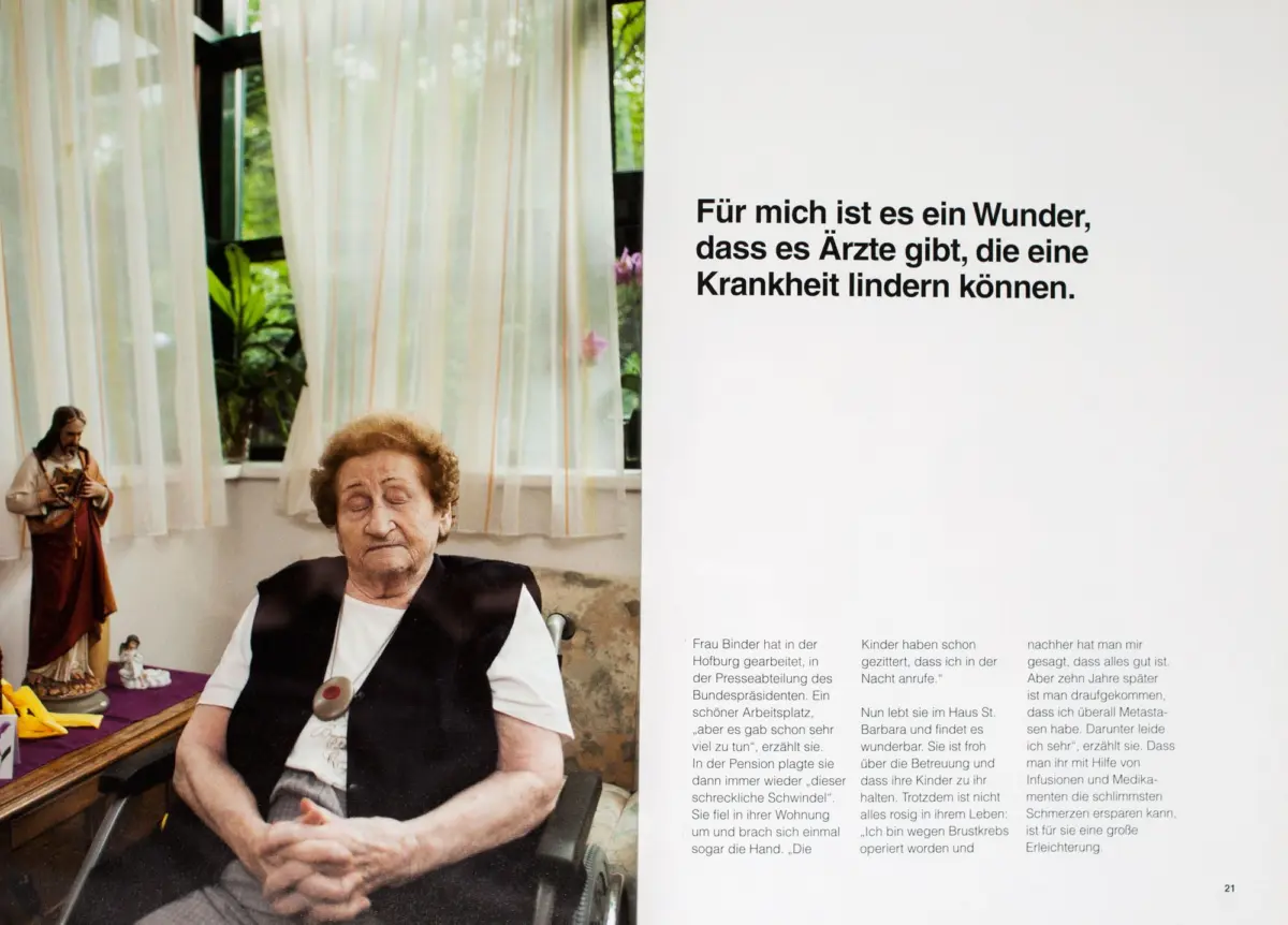 Older woman in a wheelchair with eyes closed, seated in a museum exhibit; white wall with German text and hanging curtains in the background.