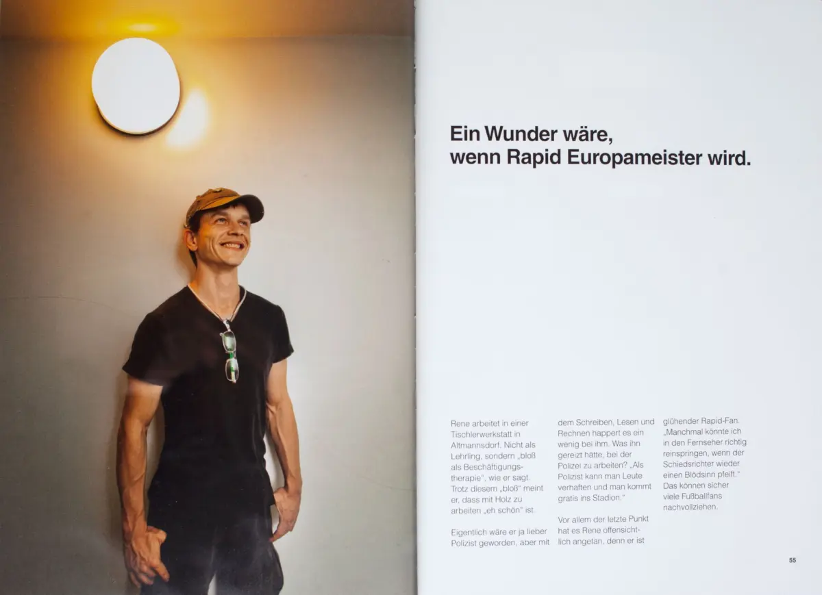 Smiling man in a black t-shirt and cap, sunglasses hanging from his shirt, stands beside a large white circular wall light in a magazine spread.