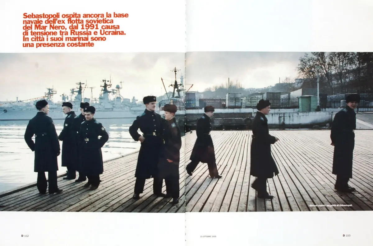 Young cadets in dark uniforms and berets stand on a wooden pier by a harbor, with warships in the background.