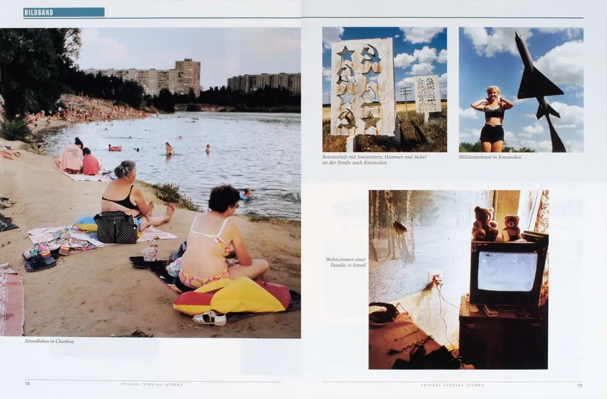 Sunbathers on a sandy river beach; people swim in the water with a skyline of apartment blocks in the background.