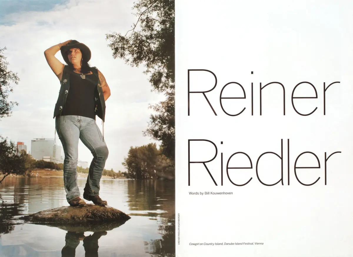 Cowgirl standing on a rock by a river, shading eyes with one hand, wearing hat and vest over jeans, with trees and city skyline in background.