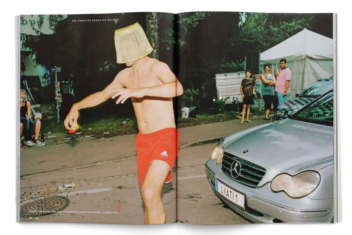 Shirtless adult man with a lampshade on his head dances outdoors; spectators near a tent and a silver car in the background.