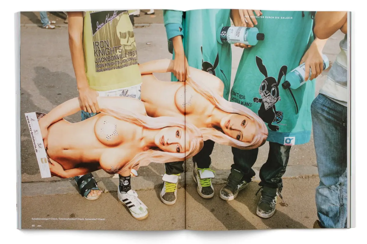 Nude model lying on side in fashion magazine spread, held open by two people outdoors; bystanders wear teal shirts.
