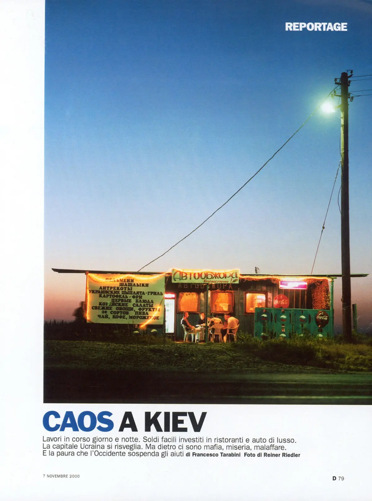 Outdoor food stall at dusk with a yellow menu banner, plastic chairs, customers, and a streetlight along a power pole against a blue sky.