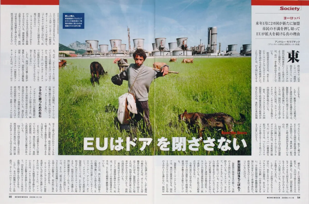 Person in gray sweatshirt stands with arms outstretched in a grassy field, cows grazing, industrial plant with cooling towers behind.