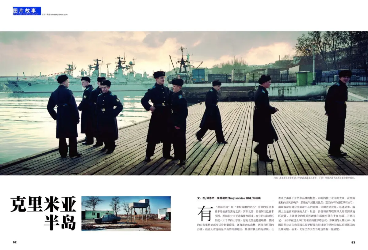 Uniformed military personnel in dark overcoats and fur hats walk along a wooden pier by a harbor, with warships in the background.