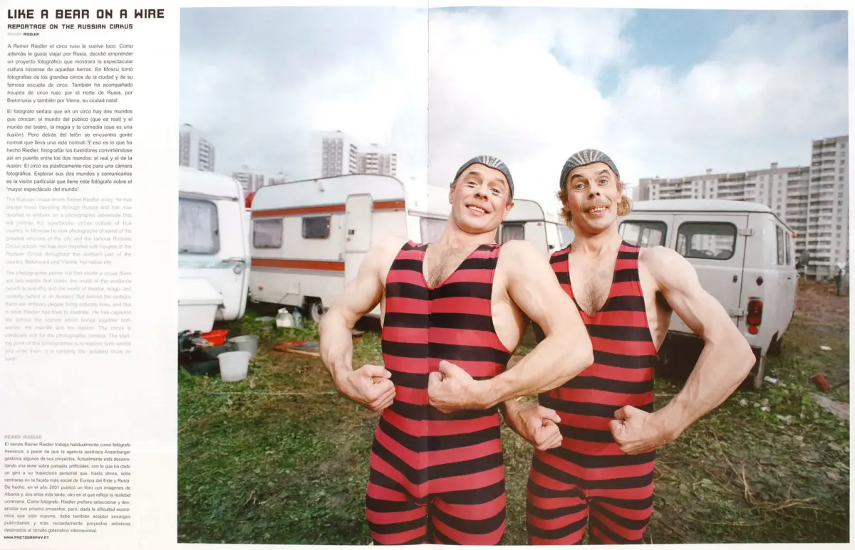 Two muscular men in red-and-black striped swimsuits and swim caps smile as they flex their arms in a yard with caravans and high-rise buildings.