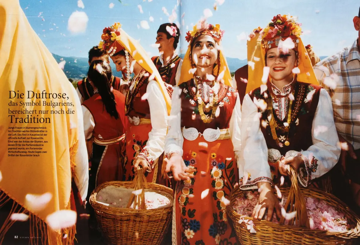 People in traditional Bulgarian folk costumes with flower crowns, carrying baskets, as confetti falls at an outdoor festival.