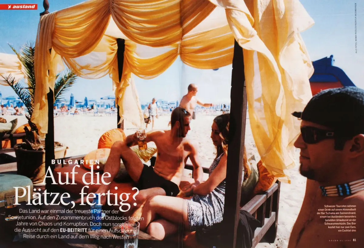 Beach scene under a flowing yellow canopy; shirtless man holds a beer while others lounge on a sunny crowded beach.