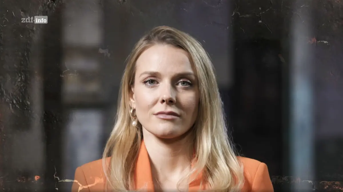 Portrait of a blonde person in an orange blazer, facing the camera in a dark studio backdrop.