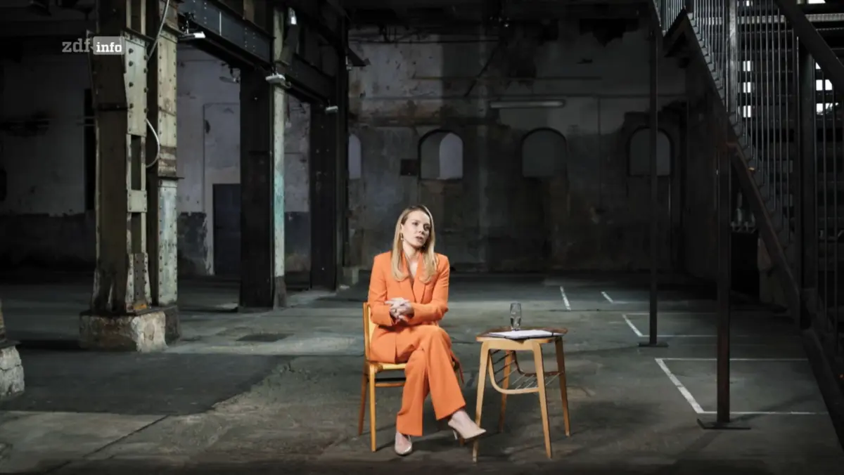 Person in orange suit sits on a wooden chair in a dim, derelict industrial warehouse; table with papers and a glass nearby.