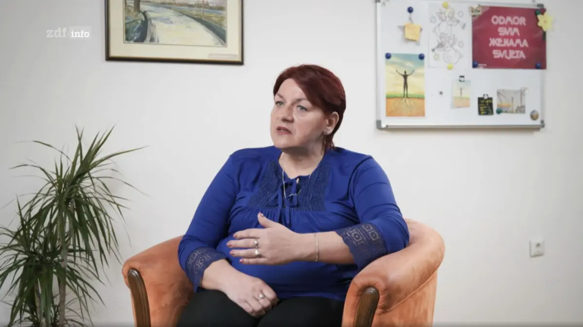 Adult person with short red hair in a blue blouse sits in an orange armchair, gesturing while talking in a small office with plants and wall art.