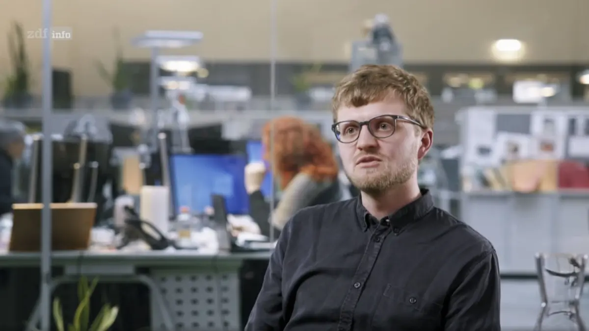 Man with glasses and stubble in a dark shirt, looking to the side in a busy open-office with desks and computers.