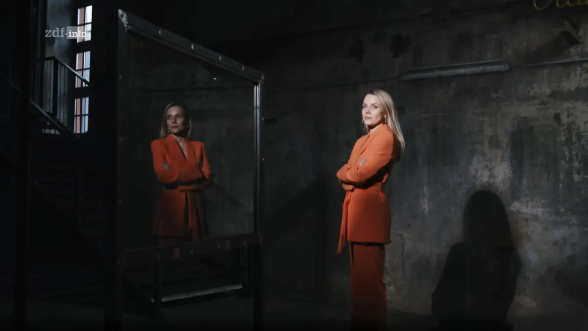 Blonde-haired person in orange pantsuit stands with arms crossed beside a metal-framed mirror, reflection visible.