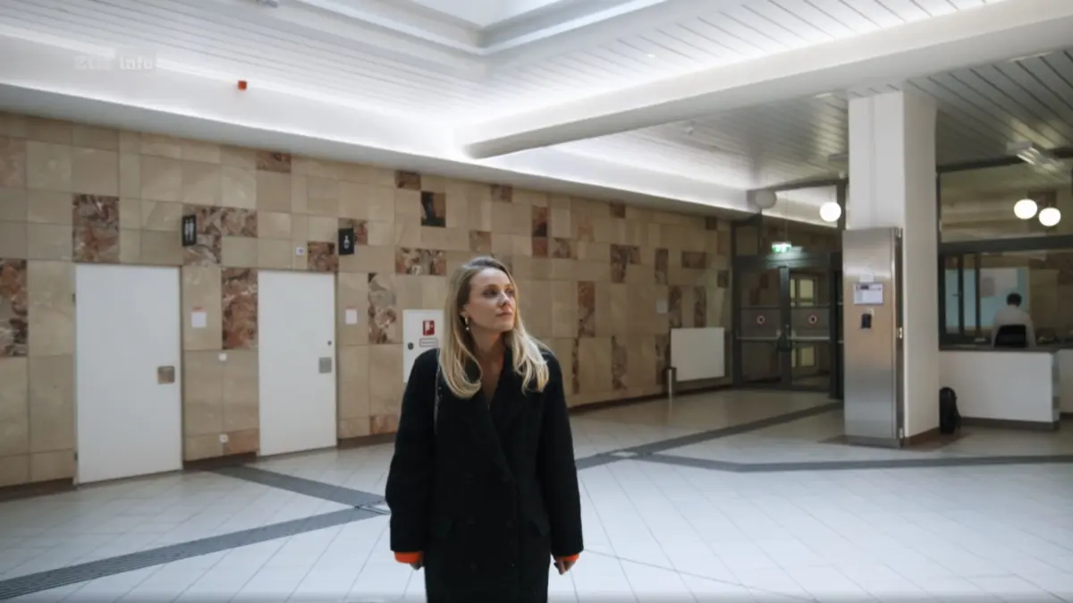Woman in a black coat stands in a spacious tiled lobby, looking to the side.