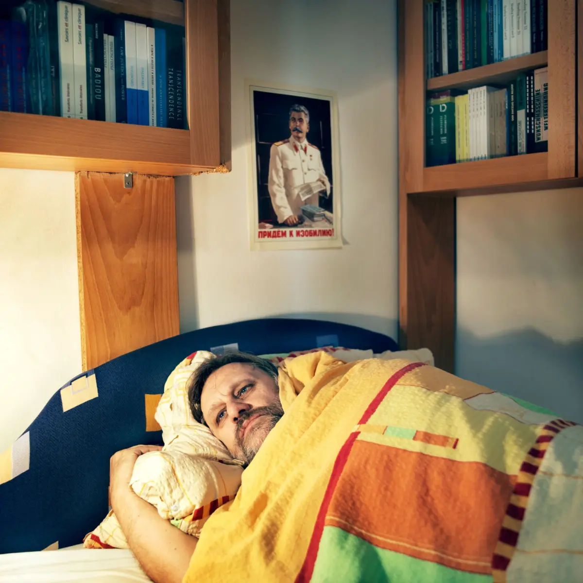 Adult person sleeping in a bed, covered by an orange-and-green striped quilt; wooden bookshelves flank the wall with a framed poster of a person in uniform.