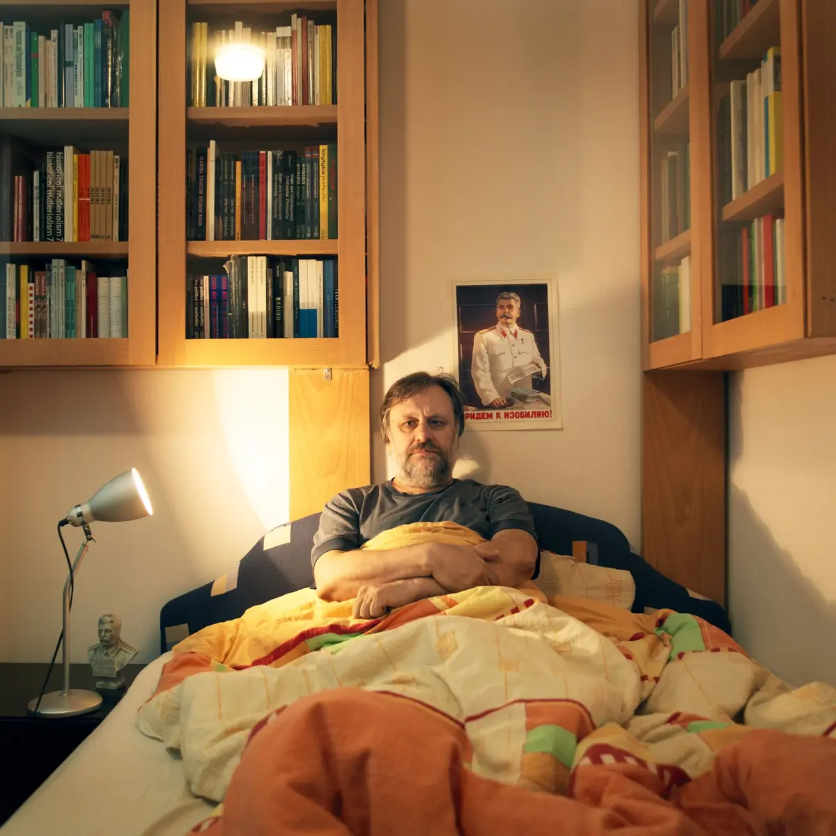 Adult person with beard sits upright on a bed, hands folded over a quilt, in a wood-paneled room with tall bookshelves and a desk lamp.