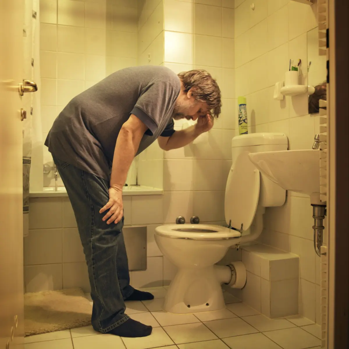 Adult person leans over toilet in a small tiled bathroom, hand to forehead.