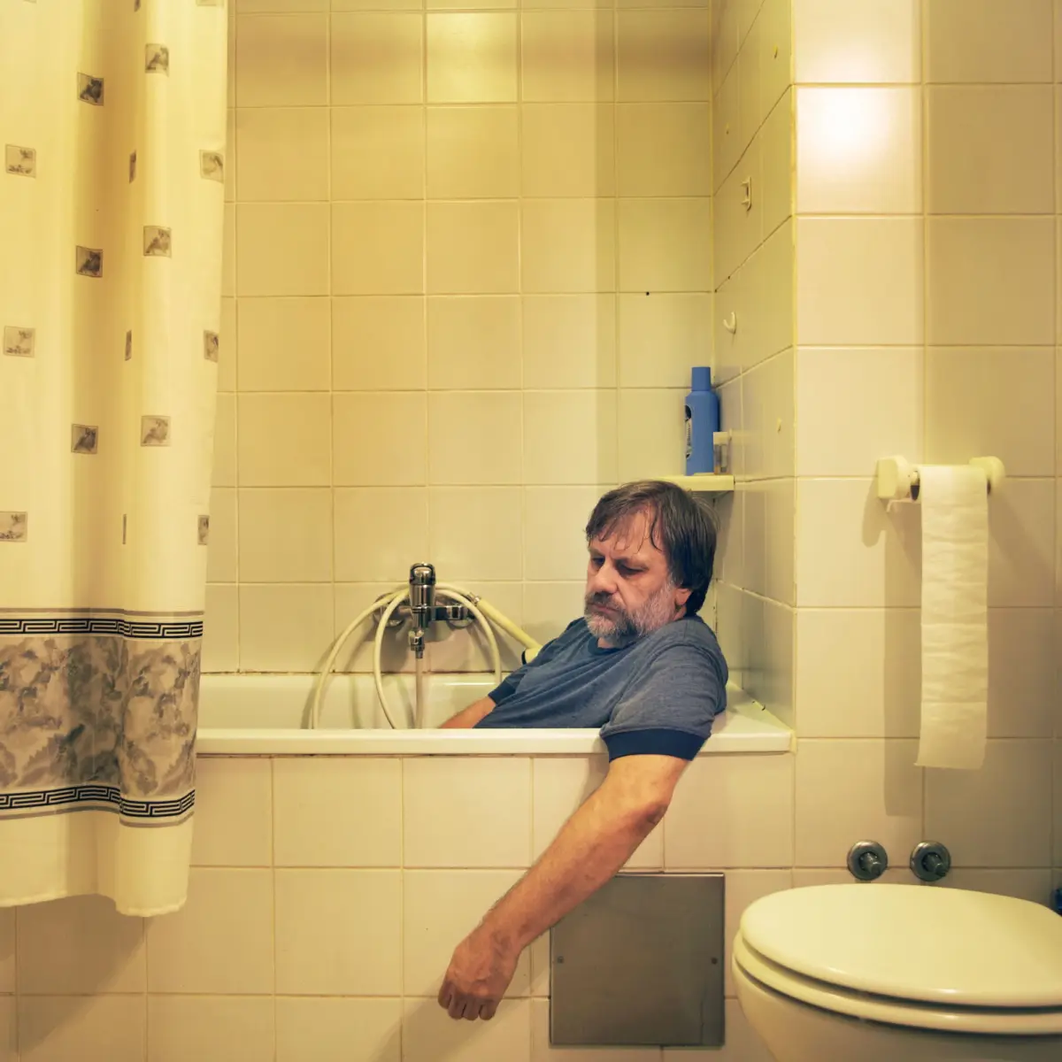 Adult person seated in a bathtub in a tiled bathroom, arm stretched toward wall; shower curtain to the left, blue bottle on a shelf, toilet nearby.