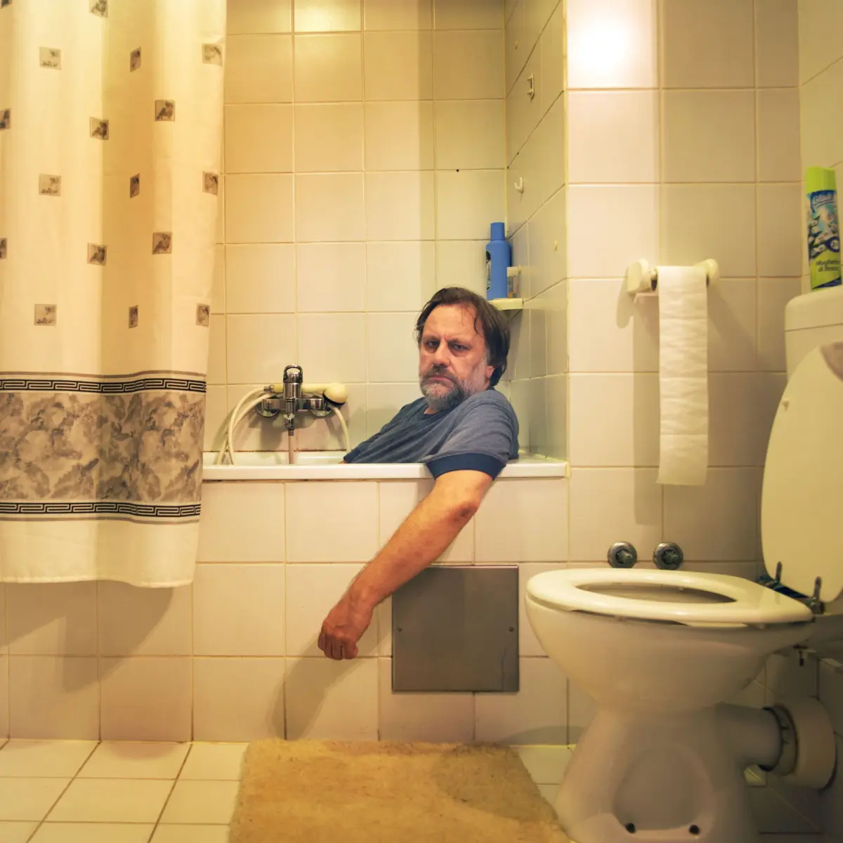 Middle-aged man sits in bathtub in tiled bathroom, arm resting on tub edge; shower curtain on left, toilet and rug visible.
