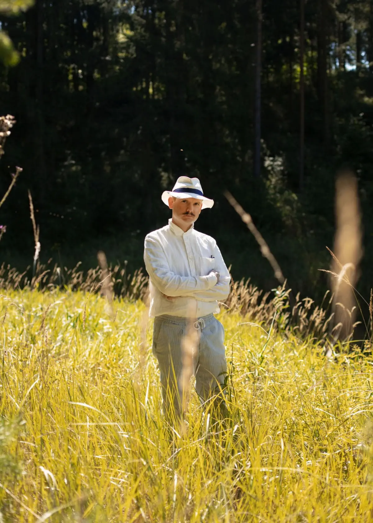Man in white shirt and Panama hat stands with arms crossed in a sunlit field of tall golden grass, with a forest behind.