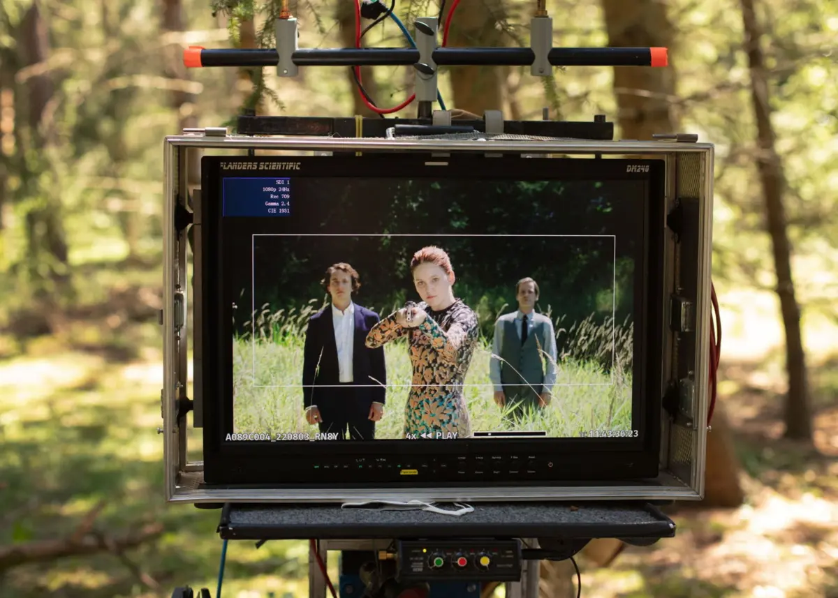 Forest film set: outdoor monitor on a rig shows three actors; woman in floral dress points a pistol at the camera.