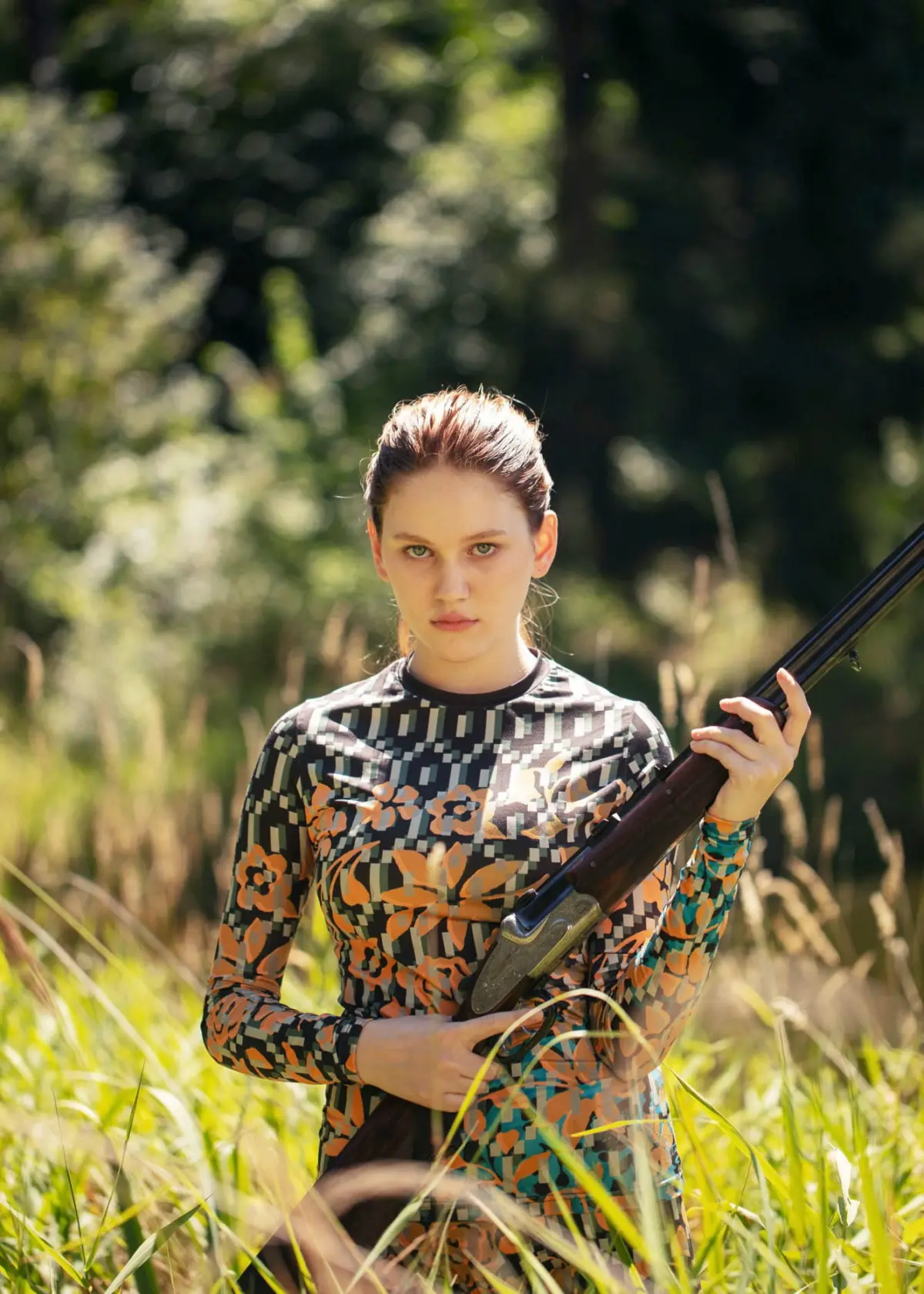 Person in patterned long-sleeve top standing in tall grass, holding a hunting rifle across their chest in a sunlit woodland clearing.