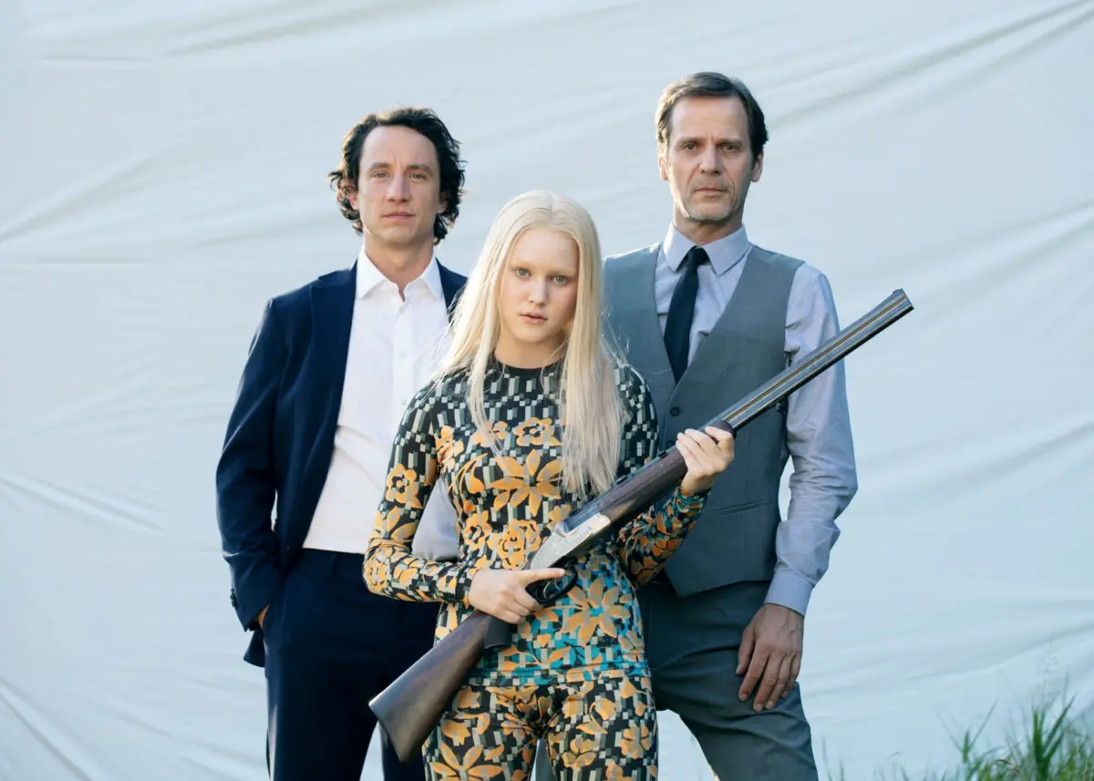 Blond-haired person in a floral dress holds a shotgun, with two men in suits behind, against a pale fabric backdrop outdoors.