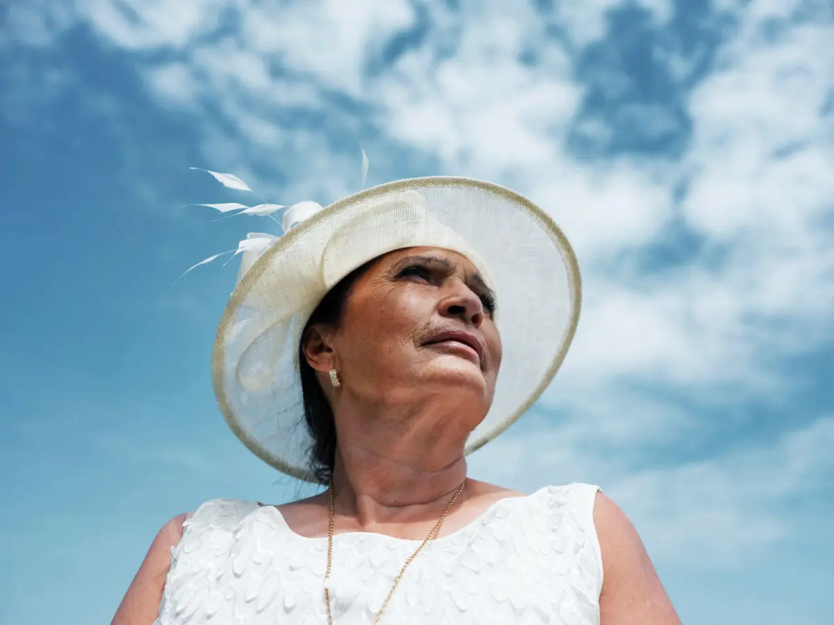 Adult wearing a white hat with feathers and a white lace dress, gazing upward against a blue sky with wispy clouds.