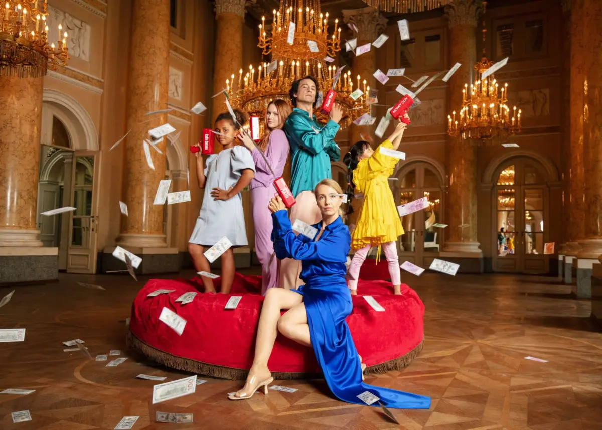 Five women in colorful outfits pose dramatically on a red circular platform as money rains in a grand marble hall.
