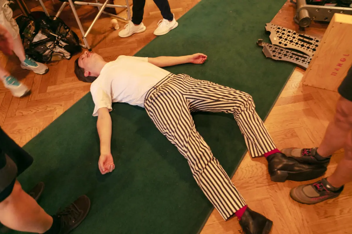 Person lying on back on green mat, arms extended, wearing white shirt and striped pants, in an indoor workshop with tools nearby.