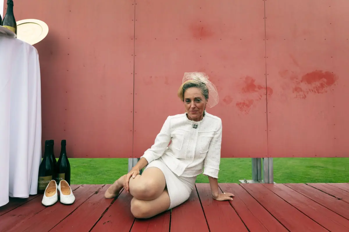 Person in a white textured suit with net fascinator sitting on a wooden deck beside a pink wall, with champagne bottles and white shoes nearby.