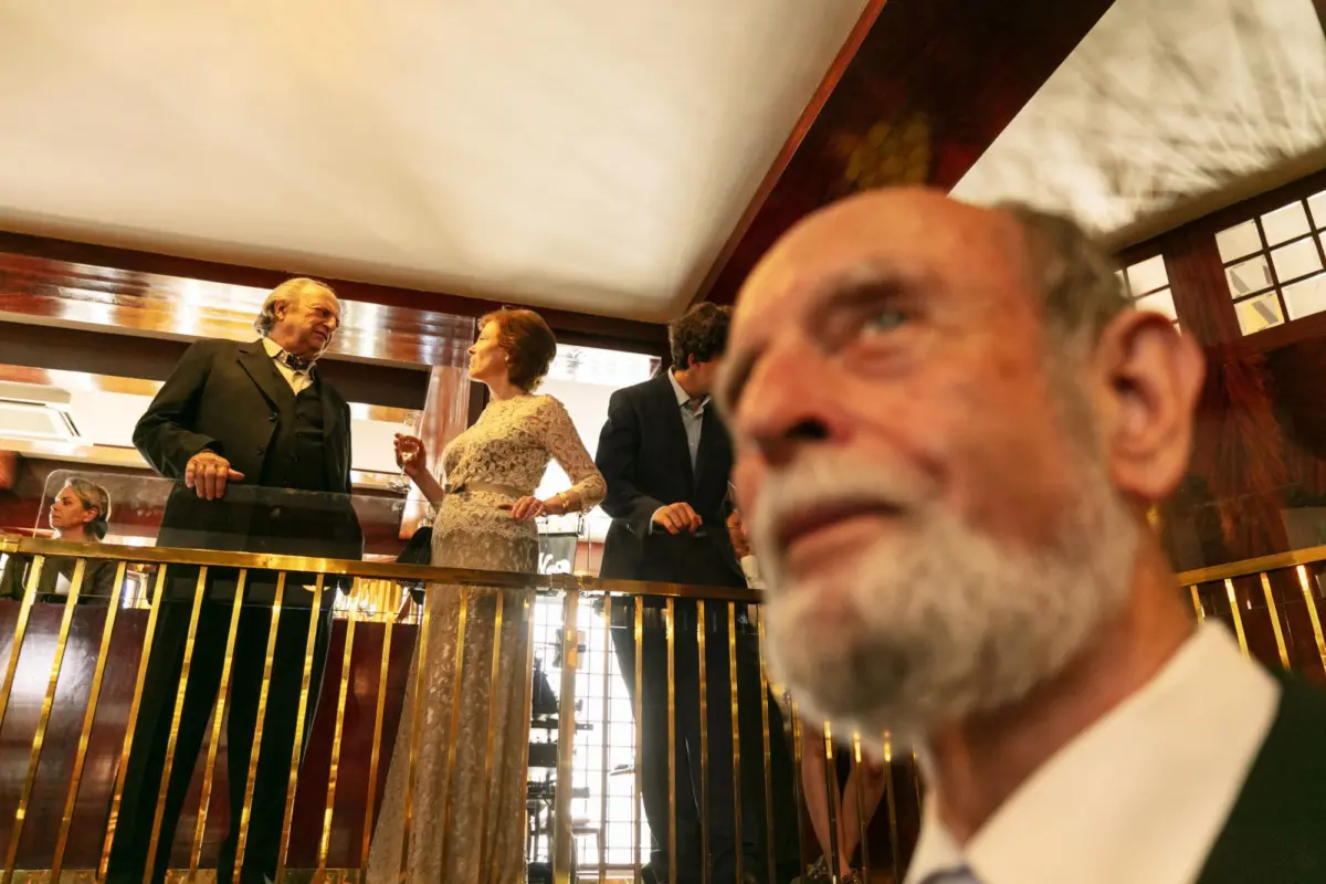 Formal event on a mezzanine balcony; suited adult talks to another in lace dress with wine; foreground bearded person partially visible.