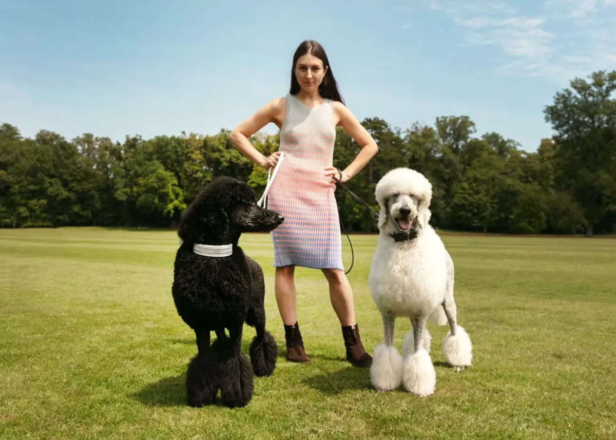 Person wearing a pastel gradient sleeveless dress stands with a black poodle and a white poodle on leashes in a sunny park.