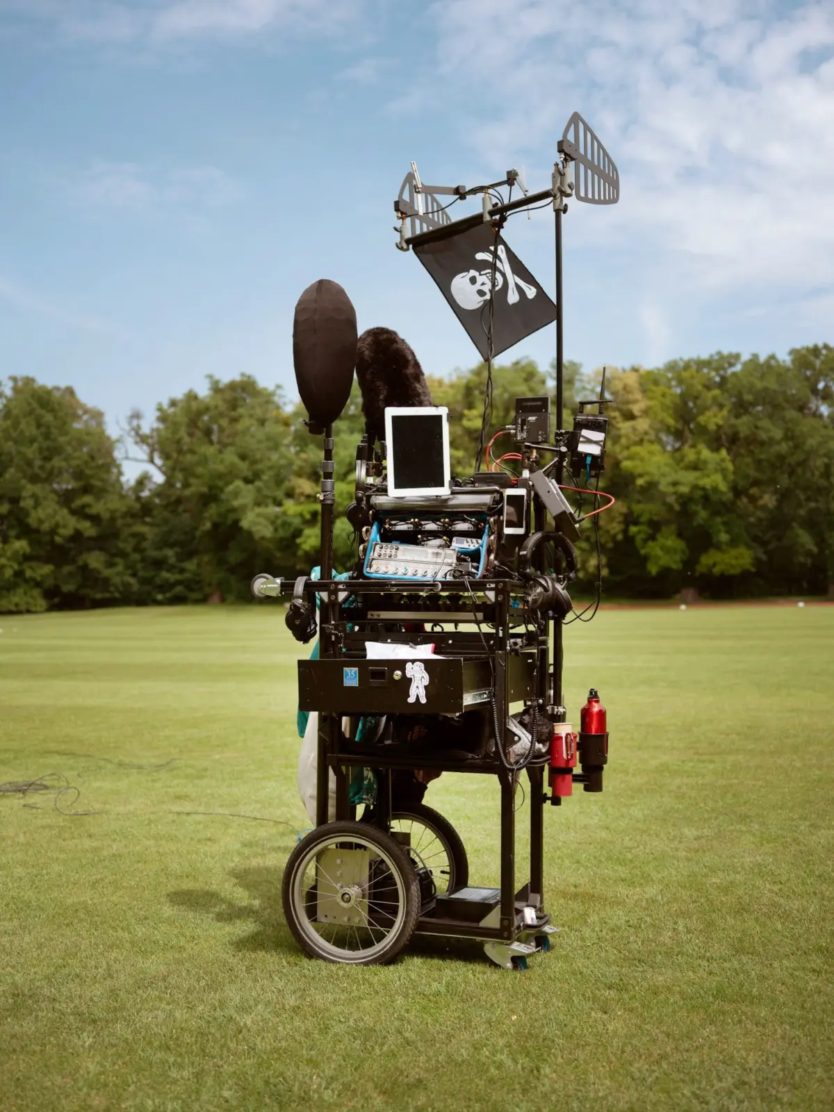 Outdoor mobile film rig on wheels with skull-and-crossbones pirate flag and monitors, on a grassy field.
