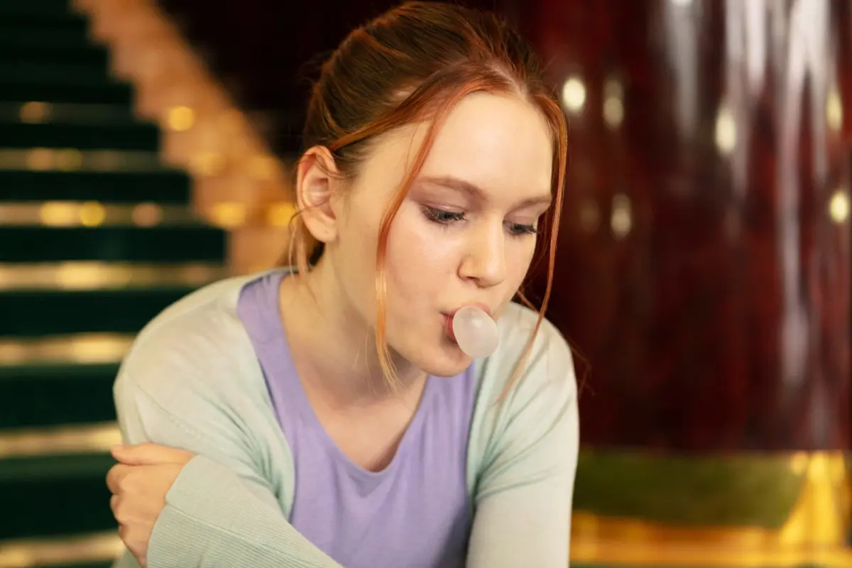 Young woman blowing a bubblegum bubble indoors near a staircase.