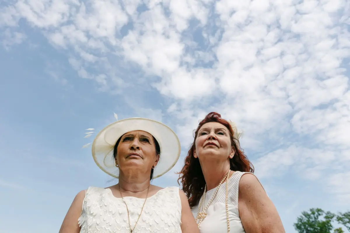 Two elderly women in white dresses, one wearing a wide-brimmed hat, stand outdoors and look upward at a cloudy blue sky.